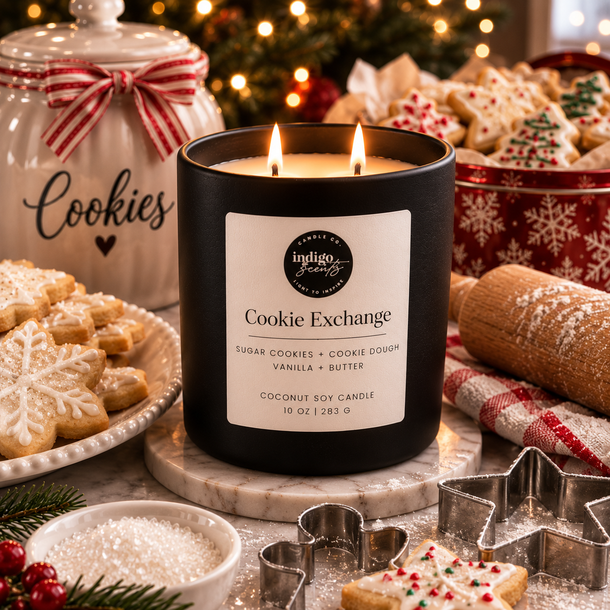 An Indigo Scents matte black candle labeled “Cookie Exchange” sits beside a cooling rack of sugar cookies, vintage cookie cutters, a rolling pin, and a tin filled with holiday treats under twinkling Christmas lights.