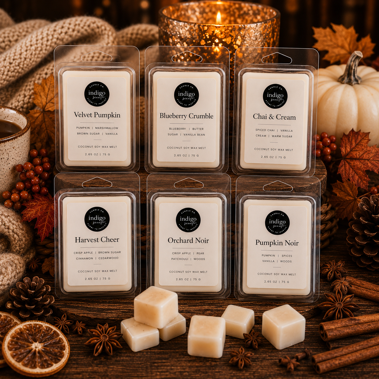 A styled product photo featuring six Indigo Scents Fall Collection wax melts in clear clamshell packaging, including Velvet Pumpkin, Blueberry Crumble, Chai & Cream, Harvest Cheer, Orchard Noir, and Pumpkin Noir. The wax melts are arranged on a rustic wooden table surrounded by pumpkins, cinnamon sticks, pinecones, autumn leaves, a cozy knit blanket, and warm candlelight.