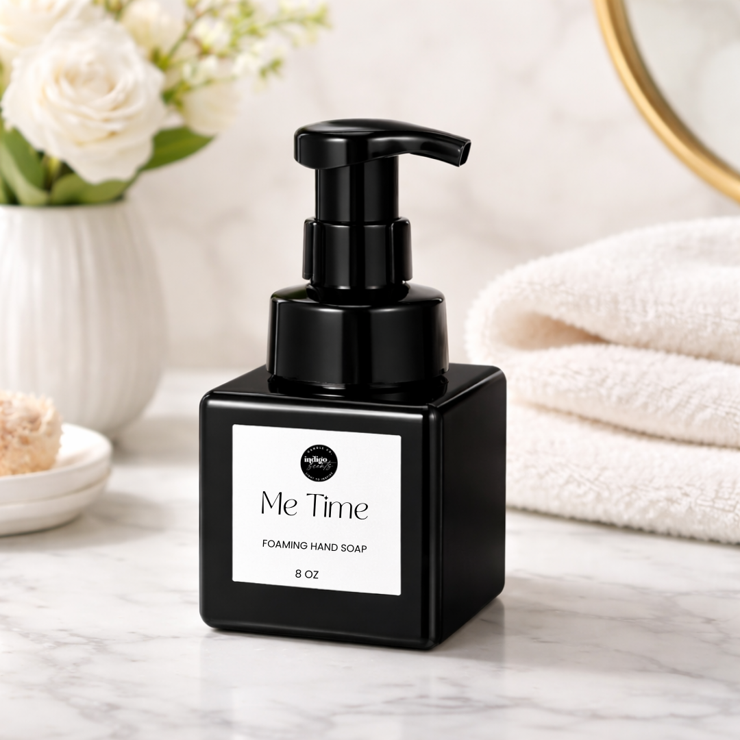 Black square bottle of Me Time foaming hand soap by Indigo Scents on a marble counter beside folded towels and white flowers.
