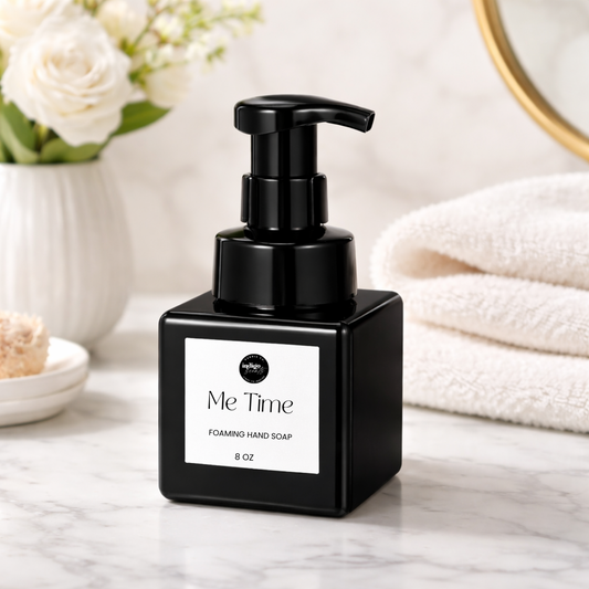 Black square bottle of Me Time foaming hand soap by Indigo Scents on a marble counter beside folded towels and white flowers.
