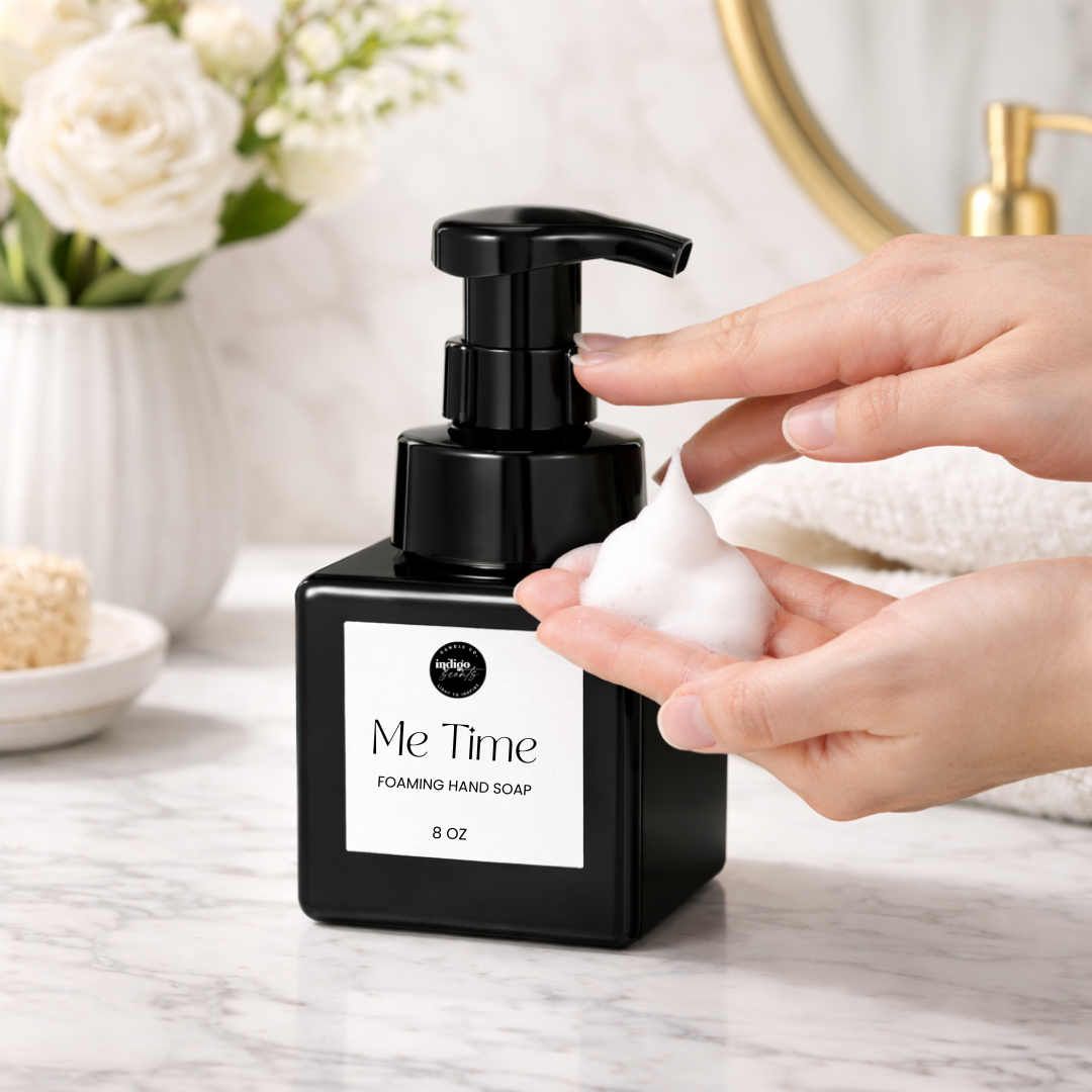 Hands dispensing foam from Indigo Scents Me Time foaming hand soap bottle on a marble bathroom counter with white flowers and towel in background.
