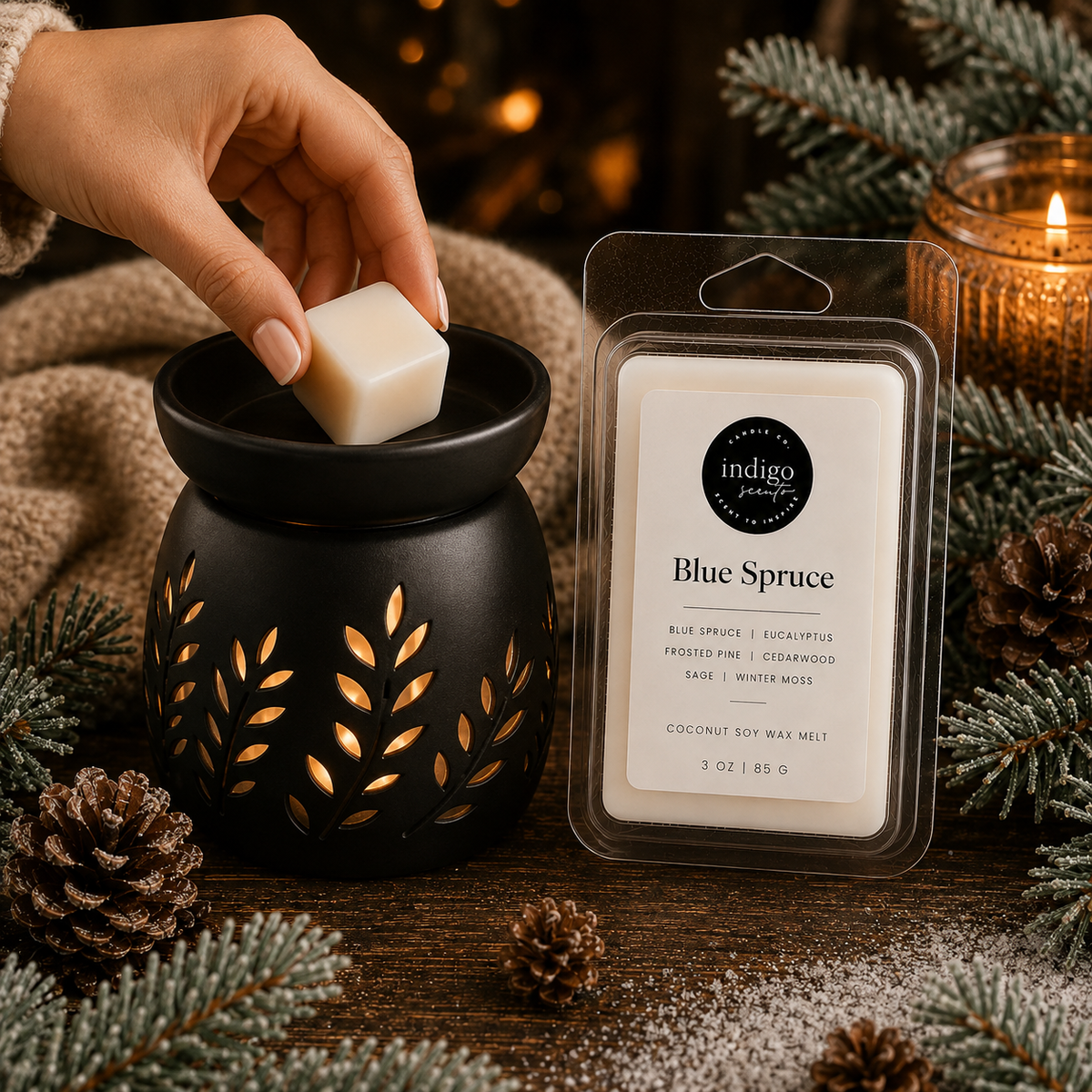 A hand holds a small off-white wax melt cube above a black ceramic wax warmer with gold leaf cutouts. Beside it is a packaged wax melt in a clear clamshell labeled 'indigo' and 'Blue Spruce.