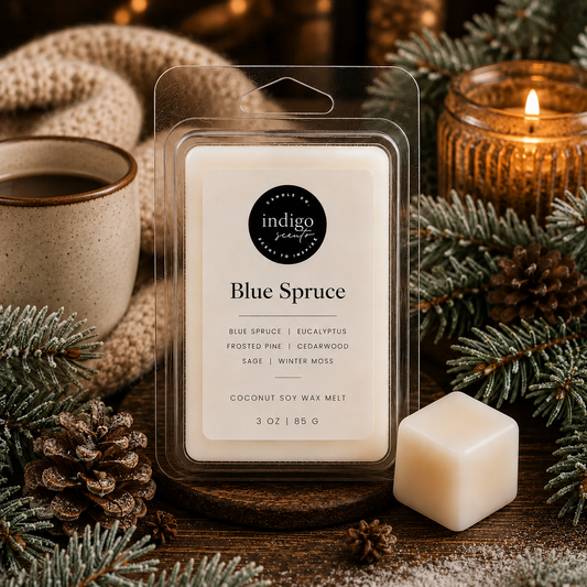 A packaged wax melt labeled 'indigo scent Blue Spruce' stands on a wooden surface, surrounded by pine branches, pinecones, a lit candle, and a small cream-colored wax cube. The product is shown in a cozy, styled holiday setting.