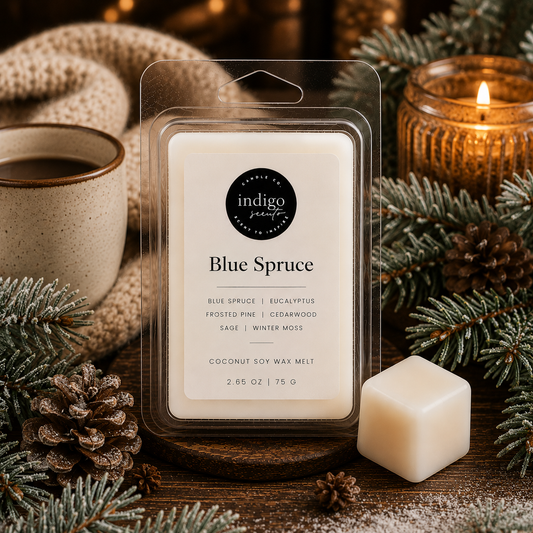 A package of Indigo Scents Blue Spruce coconut soy wax melt is displayed on a rustic wooden surface with a single wax cube next to it, surrounded by pine branches, pinecones, a cup of coffee, and a lit candle.